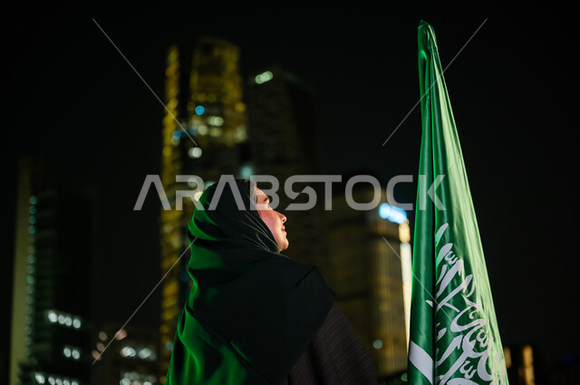  المشاركة بالمناسبات الوطنية, الوقوف بشموخ وإظهار حب الوطن, صورة مقربة من الخلف لسيدة عربية سعودية خليجية ترتدي العباءة والحجاب تنظر إلى العلم بافتخار في 11 مارس, ذكرى اليوم الوطني السعودي 23 سبتمبر, خلفية الأبراج والمباني المضيئة في وقت الليل