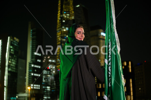 استذكار اليوم الوطني  23 سبتمبر, النظر إلى الكاميرا بإيماءات السعادة, المشاركة بالمناسبات الوطنية, صورة من الخلف لإمرأة عربية سعودية خليجية مبتسمة ترتدي العباءة والحجاب تمسك علم المملكة باحترام وتقدير, يوم العلم 11 مارس, المباني المضاءة في مدينة الرياض