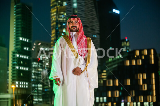 ارتداء زي المناسبات الشعبية التقليدية، الفخر والاعتزاز بالانتماء للهوية السعودية, الوقوف بثقة بإيماءات السرور والسعادة, رجل عربي خليجي سعودي يرتدي الشماغ والبشت الأبيض ينظر إلى الكاميرا, ناطحات السحاب والأبراج المضاءة ليلا في مدينة الرياض