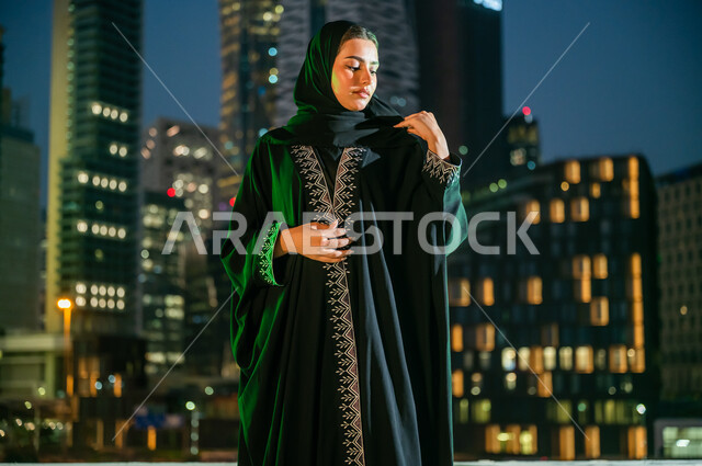 انعكاس الضوء الأخضر على مواطنة عربية خليجية سعودية تشارك في المناسبات الوطنية, النظر إلى تطريز العباءة والإعجاب بها, ارتداء الأزياء السعودية التقليدية في ذكرى اليوم الوطني 23 سبتمبر, يوم العلم السعودي 11 مارس, الأبراج والمباني المضاءة في مدينة الرياض