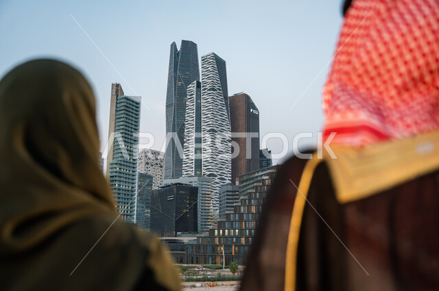 الاستمتاع بالخروج في الهواء الطلق, الوقوف أمام ناطحات السحاب في الرياض, صورة مقربة من الخلف لرجل وإمرأة عربيين سعوديين خليجيين يقضيان وقت ممتع, ارتداء الأزياء الشعبية التقليدية, خلفية الأبراج والمباني الاقتصادية, أماكن ومعالم سياحية مشهورة في السعودية