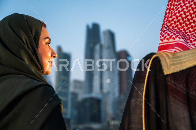 الوقوف أمام ناطحات السحاب في الرياض, الاستمتاع بالخروج في الهواء الطلق, صورة مقربة من الخلف لرجل وإمرأة عربيين سعوديين خليجيين يقضيان وقت ممتع, ارتداء الأزياء الشعبية التقليدية, خلفية الأبراج والمباني الاقتصادية, أماكن ومعالم سياحية مشهورة في السعودية