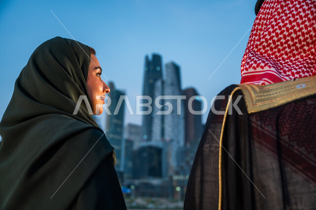 الاستمتاع بالخروج في الهواء الطلق, الوقوف أمام ناطحات السحاب في الرياض, صورة مقربة من الخلف لرجل وإمرأة عربيين سعوديين خليجيين يقضيان وقت ممتع, ارتداء الأزياء الشعبية التقليدية, خلفية الأبراج والمباني الاقتصادية, أماكن ومعالم سياحية مشهورة في السعودية