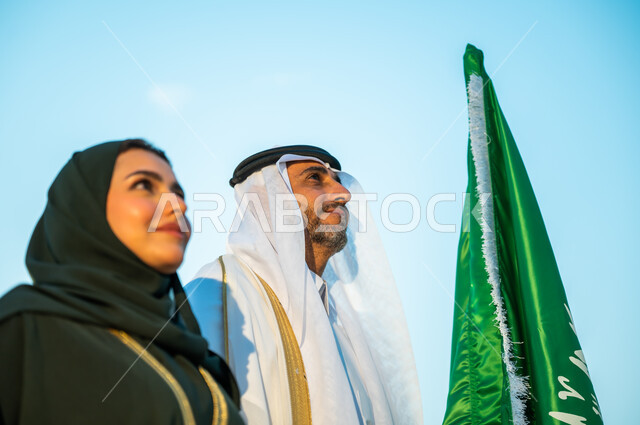ارتداء الأزياء التقليدية في ذكرى اليوم الوطني 23 سبتمبر, إحياء تاريخ توحيد المملكة, المشاركة في تنمية الوطن ورفع قدره, صورة مقربة لرجل وإمرأة عربيين سعوديين خليجيين ينظران إلى شيء ما بإيماءات السرور, الوقوف بثقة في يوم العلم السعودي 11 مارس