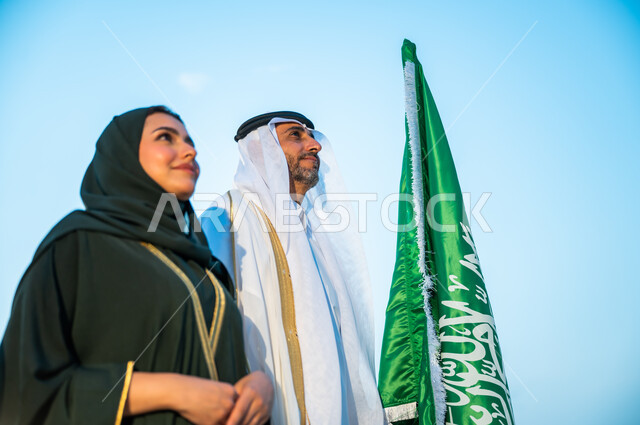 ارتداء الأزياء التقليدية في ذكرى اليوم الوطني 23 سبتمبر, إحياء تاريخ توحيد المملكة, المشاركة في تنمية الوطن ورفع قدره, صورة مقربة لرجل وإمرأة عربيين سعوديين خليجيين ينظران إلى شيء ما بإيماءات السرور, الوقوف بثقة في يوم العلم السعودي 11 مارس