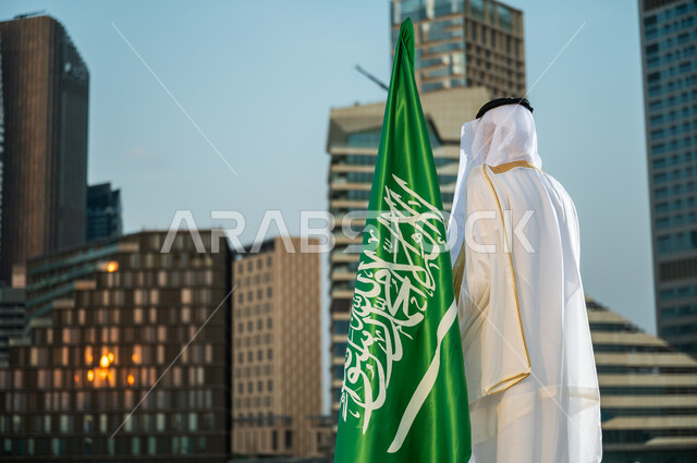 تخليد ذكرى  توحيد الوطن في 8 ربيع الأول, الوقوف لتأمل الأبراج وناطحات السحاب في وقت النهار, صورة من الخلف لرجل عربي سعودي خليجي يرتدي البشت والغترة يقف بجانب العلم,  الاحتفال باليوم الوطني, يوم العلم السعودي 11 مارس, خلفية مركز الملك عبدالله المالي