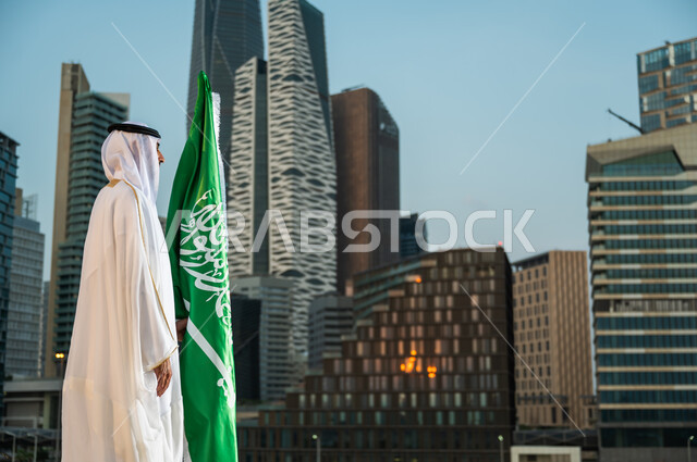 المباني والمنشآت المعمارية في مدينة الرياض, احياء ذكرى اليوم الوطني، العيد الوطني في 23 سبتمبر، الاحتفال بيوم العلم السعودي 11 مارس، صورة من الجانب لرجل عربي خليجي سعودي يرتدي الغترة والبشت يقف بجانب العلم بتقدير واحترام