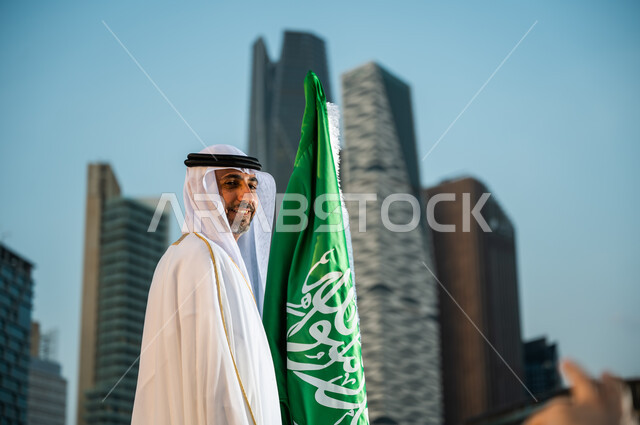 احياء ذكرى اليوم الوطني 23 سبتمبر، العيد الوطني السعودي، الاحتفال بيوم العلم 11 مارس، الوقوف بفخر وعز بإنجازات الوطن، رجل عربي خليجي سعودي مبتسم يرتدي الغترة والبشت يقف بجانب العلم بإيماءات الحب والسرور, المنشآت المعمارية في مدينة الرياض