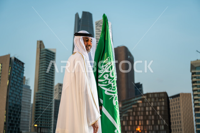 احياء ذكرى اليوم الوطني ، العيد الوطني في 23 سبتمبر، الاحتفال بيوم العلم السعودي 11 مارس، صورة من الجانب لرجل عربي خليجي سعودي مبتسم يرتدي الغترة والبشت يقف بجانب العلم بإيماءات الحب والسرور, خلفية المنشآت والمباني المعمارية في مدينة الرياض