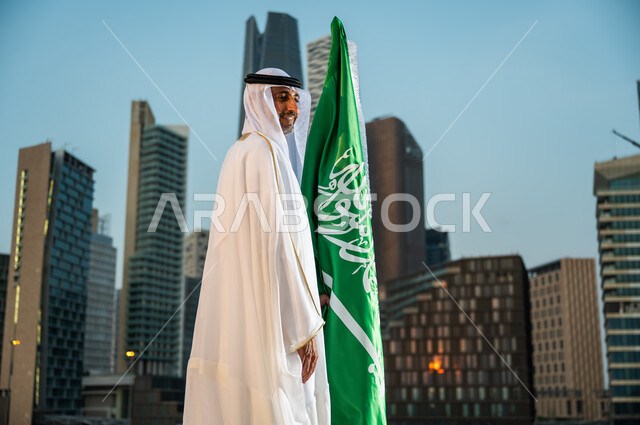 المنشآت والمباني المعمارية في مدينة الرياض, احياء ذكرى اليوم الوطني، العيد الوطني في 23 سبتمبر، الاحتفال بيوم العلم السعودي 11 مارس، صورة من الجانب لرجل عربي خليجي سعودي مبتسم يرتدي الغترة والبشت يقف بجانب العلم بإيماءات الحب والسرور