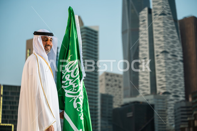 الاحتفال بيوم العلم السعودي 11 مارس, احياء ذكرى اليوم الوطني، العيد الوطني في 23 سبتمبر، صورة من الجانب لرجل عربي خليجي سعودي مبتسم يرتدي الغترة والبشت يقف بجانب العلم بإيماءات الحب والسرور, خلفية المنشآت والمباني المعمارية في مدينة الرياض