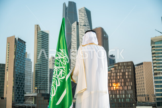 Celebrating National Day, September 23, in front of the Caved Towers, attention to the financial center in Riyadh, a picture from behind, of a Saudi Gulf Arab man wearing the traditional Saudi bisht and ghutra, looking at the financial towers, Science Day, March 11, pride in the urban development of economic facilities