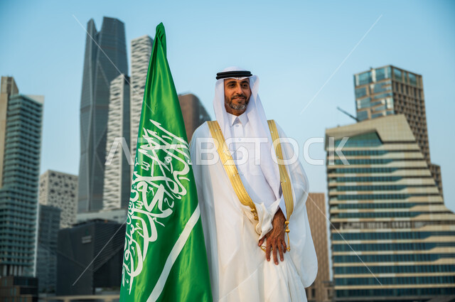 النظر إلى شيء ما بإيماءات السرور, الاهتمام بالمركز المالي في الرياض, رجل عربي سعودي خليجي يرتدي البشت والغترة التقليدية يمسك العلم باحترام وتقدير, الاحتفال باليوم الوطني 8 ربيع الأول, ذكرى يوم العلم 11 مارس, النمو الاقتصادي والمالي في المملكة