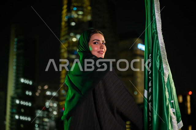 المباني المضاءة في وقت الليل في مدينة الرياض, ايماءات السعادة والسرور بيوم العلم 11 مارس, صورة مقربة من الخلف لإمرأة عربية سعودية خليجية مبتسمة ترتدي العباءة والحجاب تمسك علم المملكة باحترام وتقدير, استذكار اليوم الوطني 23 سبتمبر