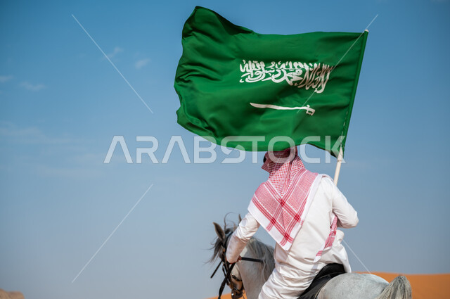 الفخر بإمتلاك الهوية السعودية، أنشطة وفعاليات ركوب الخيول في الصحراء، صورة من الخلف لخيال عربي خليجي سعودي يمتطي فرس عربي أصيل ويرفع علم السعودية، العيد الوطني السعودي، اليوم الوطني لتوحيد المملكة 23 سبتمبر، يوم العلم 11 مارس، توارث شغف الجياد من الأجداد