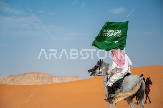 العيد الوطني السعودي ، شغف الفروسية وممارسة هواية ركوب الخيل في الصحراء، صور من الخلف لفارس عربي خليجي سعودي يمتطي فرس عربي أصيل ويرفع علم السعودية، اليوم الوطني السعودي 23 سبتمبر، يوم العلم 11 مارس، إظهار مشاعر الاعتزاز بالوطن، نشر حب الخيل بين ابناء الوطن