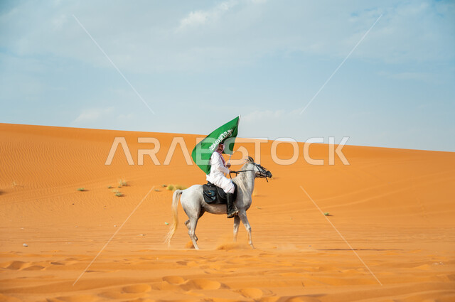 Participating in the development of the nation and raising its value, a picture from the side of a Saudi Arabian Gulf horseman riding an Arabian horse and raising the flag of Saudi Arabia, the anniversary of the National Day for the Unification of the Kingdom, the Saudi National Day 8 Rabi’ al-Awwal, Science Day March 11, the passion for equestrianism and practicing the hobby of horse riding in the desert