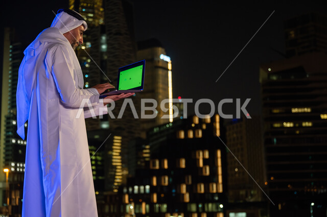 عقد صفقات عمل مع رجال اعمال, صورة مقربة من الخلف لرجل عربي سعودي خليجي يرتدي الزي التقليدي يقوم باستخدام جهاز الحاسوب المحمول بخلفية شاشه خضراء كروما, خلفية الأبراج وناطحات السحاب المضيئة ليلا في مدينة الرياض، استخدام التقنيات الحديثة المتطور في العمل