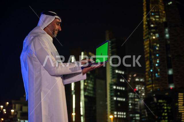 تصفح مواقع التواصل الاجتماعي، صورة مقربة من الجانب لرجل عربي سعودي خليجي يرتدي الزي التقليدي يقوم باستخدام جهاز الحاسوب المحمول بخلفية شاشه خضراء كروما, خلفية الأبراج وناطحات السحاب المضيئة ليلا في مدينة الرياض، استخدام التقنيات الحديثة المتطور في العمل