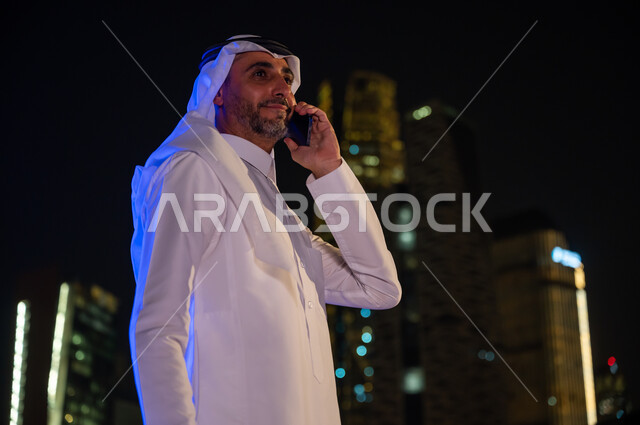 عقد صفقات عمل عن طريق التقنيات الحديثة ,معالم سياحية مشهورة، إجراء مكالمة صوتية هاتفيه، صورة مقربة لرجل عربي خليجي سعودي يرتدي الزي السعودي التقليدي يحمل بيده الهاتف المحمول في وقت الليل، خلفية الأبراج وناطحات السحاب المضيئة ليلا في مدينة الرياض