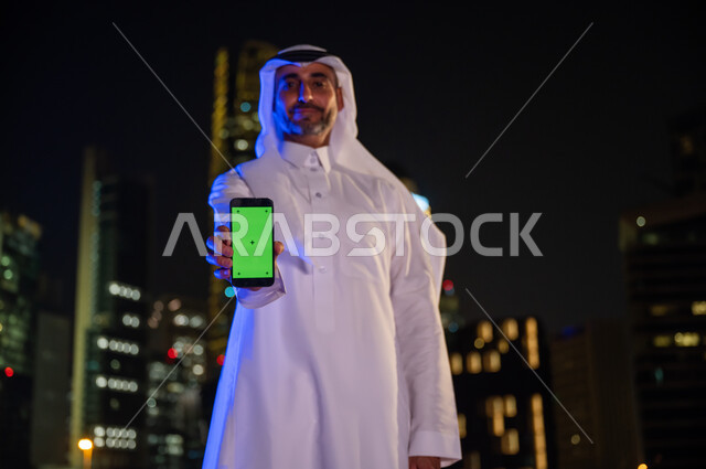 معالم مشهورة سياحية, الوقوف بثقة واعتزاز ،رجل أعمال عربي سعودي خليجي مبتسم يرتدي الزي السعودي التقليدي يقوم بعرض شاشه خضراء عبر الهاتف المحمول و ينظر إلى الكاميرا, خلفية الأبراج وناطحات السحاب المضيئة ليلا في مدينة الرياض, كروما