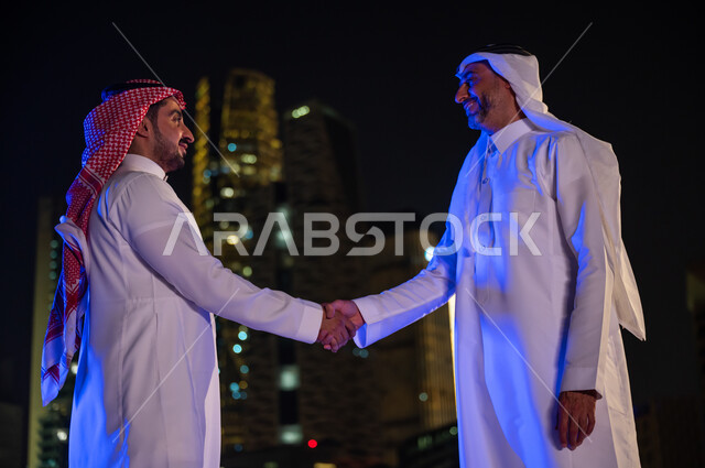 عقد صفقات عمل جديدة ناجحة بين رجال الأعمال، تعارف وصداقات وبناء علاقات اجتماعية، الخروج في الهواء الطلق، رجلان عربيان خليجيان سعوديان مبتسمان يرتديان الزي التقليدي السعودي يقومان بمصافحة بعضهما، خلفية أبراج ومباني مدينة الرياض المضيئة ليلا