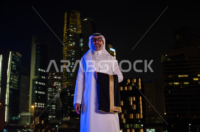 الوقوف باستقامة و ثقه  واعتزاز بالدولة السعودية والنظر إلى الكاميرا، ايماءات تدل على السعادة والسرور، رجل عربي خليجي سعودي يرتدي الثوب التقليدي والنظارة الطبيه ويحمل بيده البشت، معالم سياحيه مشهورة، خلفيه أبراج مدينة الرياض المضيئة ليلا
