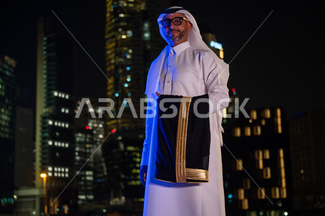 الوقوف بثقه واعتزاز بالدولة السعودية والنظر إلى الكاميرا، ايماءات تدل على السعادة والسرور، رجل عربي خليجي سعودي يرتدي الثوب التقليدي والنظارة الطبيه ويحمل بيده البشت، معالم سياحيه مشهورة، خلفيه أبراج مدينة الرياض المضيئة ليلا
