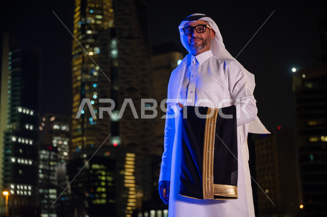 ايماءات تدل على السعادة والسرور، رجل عربي خليجي سعودي يرتدي الثوب التقليدي والنظارة الطبيه ويحمل بيده  البشت، الوقوف بثقه واعتزاز بالدولة السعودية والنظر إلى الكاميرا، معالم سياحيه مشهورة، خلفيه أبراج مدينة الرياض المضيئة ليلا