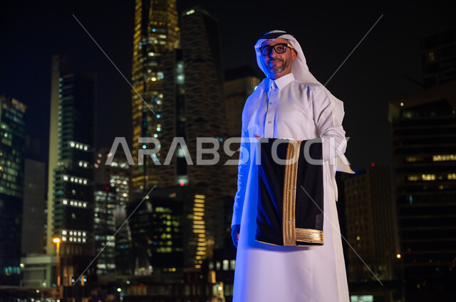 معالم سياحيه مشهورة، ايماءات تدل على السعادة والسرور، رجل عربي خليجي سعودي يرتدي الثوب التقليدي والنظارة الطبيه ويحمل بيده  البشت، الوقوف بثقه واعتزاز بالدولة السعودية والنظر إلى الكاميرا، خلفيه أبراج مدينة الرياض المضيئة ليلا