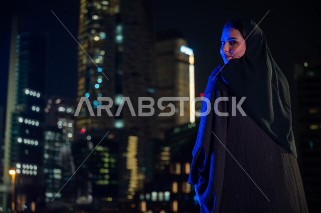 Gestures of pride in growth and urban progress and looking at the camera, a close-up of a veiled Saudi Arabian Gulf woman wearing the Gulf abaya and standing in front of the luminous towers of the city of Riyadh at night, standing with confidence and prid