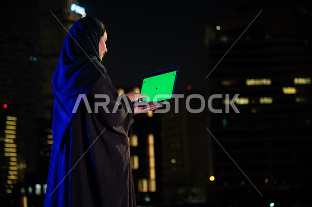 Surfing social networking sites, spending quality time using modern devices and technologies, enjoying the outdoors outside, a close-up image from the back of a Saudi woman wearing a Gulf abaya holding a computer with a blank green screen, the background 