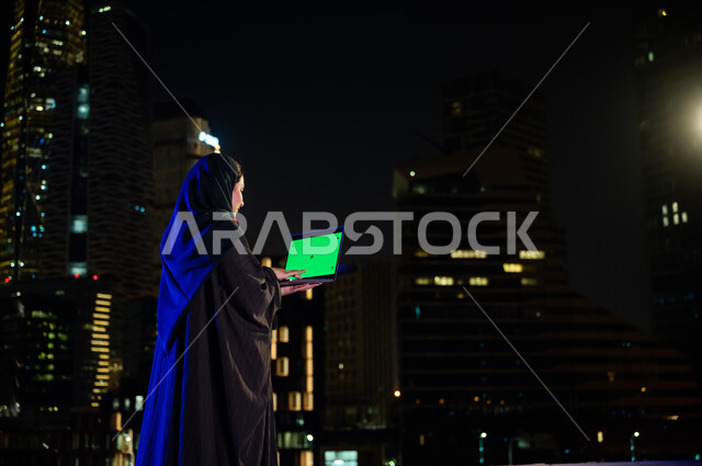 قضاء وقت ممتع باستخدام الأجهزة والتقنيات الحديثة, الاستمتاع بالهواء الطلق في الخارج, صورة من الخلف لإمرأة عربية سعودية ترتدي العباءة الخليجيه تحمل جهاز الحاسوب بشاشة خضراء فارغة, تصفح مواقع التواصل الاجتماعي، خلفية الابراج وناطحات السحاب المضيئة، كروما 