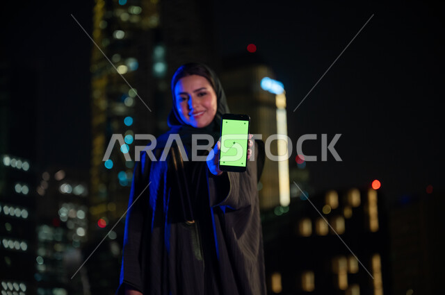 ايماءات السعادة والسرور والنظر إلى الكاميرا ،عرض شاشه خضراء عبر الهاتف المحمول ،صورة مقربة لإمرأة خليجيه سعوديه محجبه مبتسمة ترتدي العباءه الخليجيه تقف امام أبراج مدينة الرياض المضيئة ليلا، معالم سياحيه مشهورة، استخدام التقنيات الحديثة المتطورة، كروما