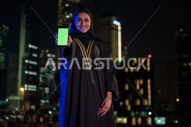 Viewing a green screen via a mobile phone, a smiling, veiled Saudi Arabian Gulf woman wearing the Gulf abaya standing in front of the illuminated towers of the city of Riyadh at night, gestures of happiness and pleasure and looking at the camera, famous t