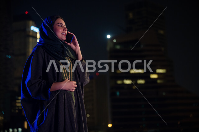 خلفية أبراج مدينة الرياض المضيئة ليلا، تبادل الأحاديث المسليه عبر الجوال، التواصل و الاطمئنان على الأصدقاء والأقارب ،امرأة عربيه خليجيه سعوديه محجبه مبتسمة ترتدي العباءه الخليجيه تقوم بإجراء مكالمة هاتفيه صوتية عبر الهاتف المحمول