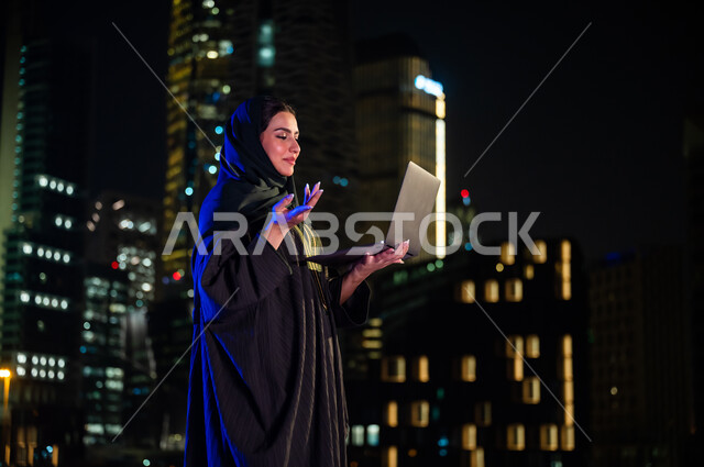 ايماءات السعادة والسؤال عن شيء ما، إجراء مكالمة فيديو عبر جهاز الحاسوب المحمول ، إلقاء التحية والسلام، استخدام الأجهزة والتقنيات الحديثة, صورة من الجانب لإمرأة سعودية محجبه ترتدي العباءة الخليجيه تقف أمام الابراج وناطحات السحاب المضيئة، مفهومم صلة الرحم