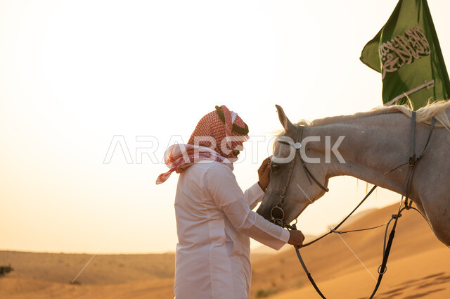 National Day for the Unification of the Country 8 Rabi’ al-Awwal, Celebrating Saudi National Day with Arabian horses, taking care of the horse at sunset in the desert, a close-up photo of a Saudi Arabian Gulf horseman wearing a shemagh and traditional dress holding a purebred Arabian horse, commemorating Flag Day March 11