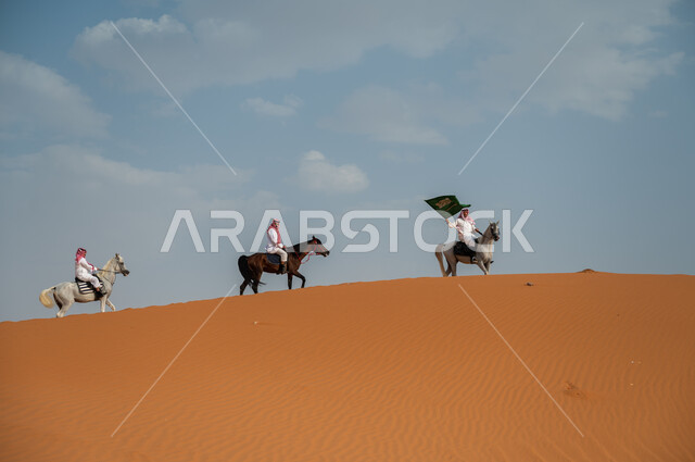 مشاركة فعاليات ركوب الخيل في العيد الوطني، هواية امتطاء الجياد في الصحراء، إحياء ذكرى اليوم الوطني 8 ربيع الأول، الاحتفال بيوم العلم السعودي 11 مارس، مجموعة رجال عرب خليجيون سعوديون يتسابقون على ظهر الاحصنة ويرفعون علم المملكة، قوة وسرعة الخيل السعودي