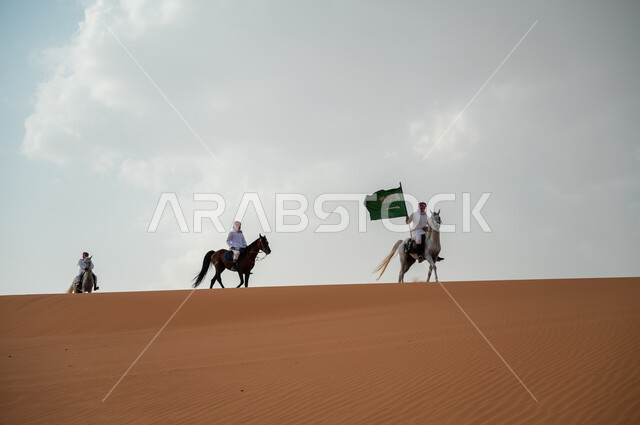 هواية امتطاء الجياد في الصحراء، إحياء ذكرى اليوم الوطني 8 ربيع الأول، مشاركة فعاليات ركوب الخيل في العيد الوطني، الاحتفال بيوم العلم السعودي 11 مارس، مجموعة رجال عرب خليجيون سعوديون يتسابقون على ظهر الاحصنة ويرفعون علم المملكة، قوة وسرعة الخيل السعودي