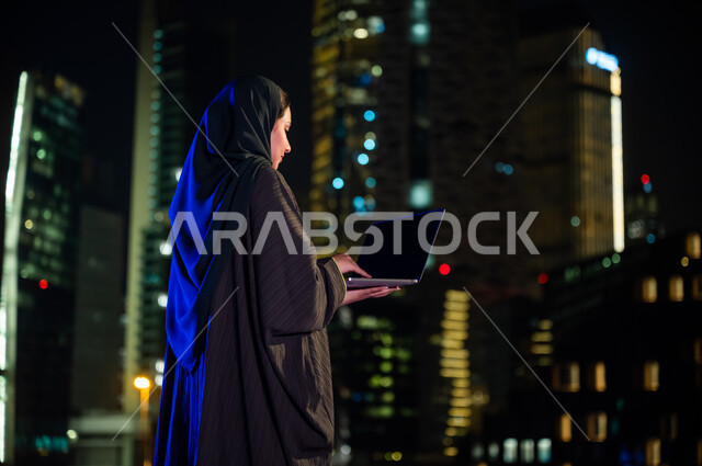 استخدام الأجهزة والتقنيات الحديثة, التسويق الالكتروني في الهواء الطلق, صورة من الخلف لإمرأة سعودية محجبه ترتدي العباءة الخليجيه تقف أمام الابراج وناطحات السحاب المضيئة، عرض شاشة سوداء فارغه عبر جهاز الحاسوب المحمول , تصفح مواقع التواصل الاجتماعي
