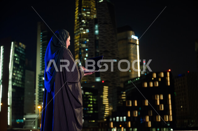 التسويق الالكتروني في الهواء الطلق, استخدام الأجهزة والتقنيات الحديثة, صورة من الخلف لإمرأة سعودية محجبه ترتدي العباءة الخليجيه تقف أمام الابراج وناطحات السحاب المضيئة، عرض شاشة سوداء فارغه عبر جهاز الحاسوب المحمول , تصفح مواقع التواصل الاجتماعي