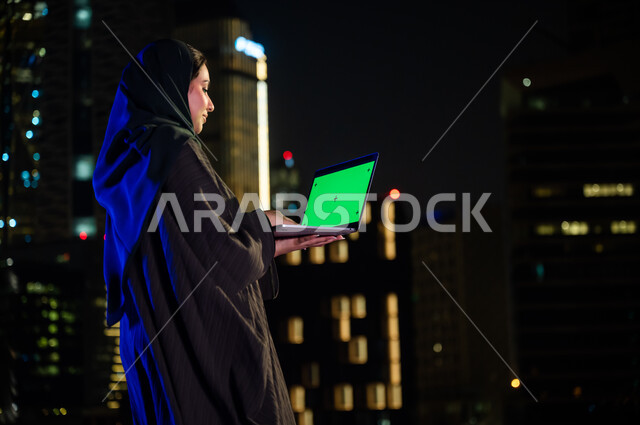 المحادثة و الدردشات مع الأصدقاء، تصفح مواقع التواصل الاجتماعي، استخدام الأجهزة والتقنيات الحديثة, صورة مقربة من الخلف لإمرأة سعودية ترتدي العباءة الخليجيه تحمل جهاز الحاسوب بشاشة خضراء فارغة, خلفية الابراج وناطحات السحاب المضيئة، كروما