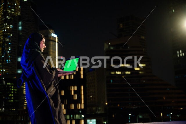 العمل عن بعد في الهواء الطلق ,تصفح مواقع التواصل الاجتماعي، استخدام الأجهزة والتقنيات الحديثة, صورة من الخلف لإمرأة سعودية ترتدي العباءة الخليجيه تحمل جهاز الحاسوب بشاشة خضراء فارغة, خلفية الابراج وناطحات السحاب المضيئة، كروما