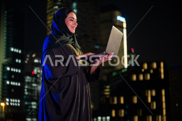 ايماءات السعادة والسرور، العمل عن بعد عبر جهاز الحاسوب المحمول, متابعه آخر أخبار السوشال ميديا، استخدام الأجهزة والتقنيات الحديثة, صورة مقربة من الجانب لإمرأة سعودية محجبه ترتدي العباءة الخليجيه تقف أمام الابراج وناطحات السحاب المضيئة