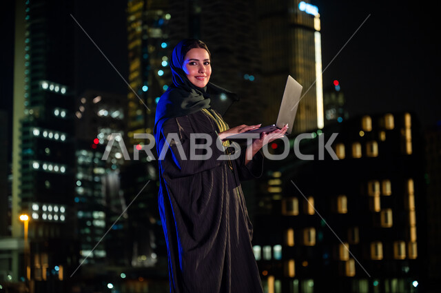 Remote work via a computer, the use of modern devices and technologies, electronic marketing in the open air, a side image of a veiled Saudi woman wearing the Gulf abaya, standing in front of the towers and bright skyscrapers and looking at the camera, br