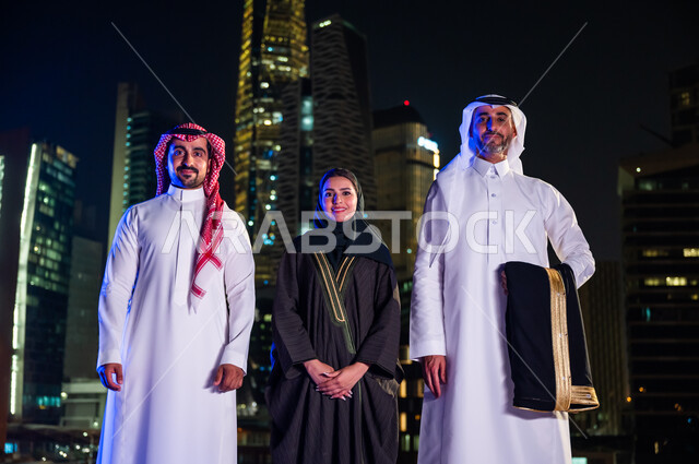 Gestures of happiness with progress and urban development, standing with confidence and pride in the Saudi state and looking at the camera, smiling Saudi Arab citizens wearing traditional dress and standing in front of the illuminated towers and skyscrape