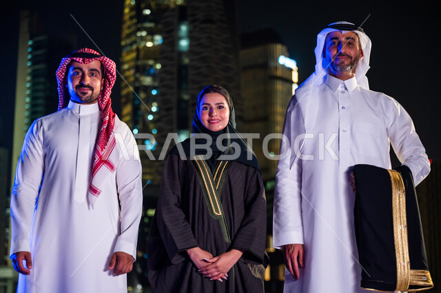 Standing upright, confident, and proud of the Saudi state and looking at the camera. Smiling Saudi Arab citizens dressed in traditional costume in front of the luminous towers and skyscrapers at night in the city of Riyadh. Gestures of happiness with prog