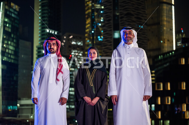 فخر أبناء المملكة بتطورها العمراني، الوقوف باستقامة وثقه واعتزاز بالدولة السعودية والنظر إلى الكاميرا، مواطنون عرب سعوديون مبتسمون يرتدون الزي التقليدي أمام أبراج وناطحات السحاب المضيئة ليلا في مدينة الرياض، ايماءات السعادة والسرور بالتقدم والنمو