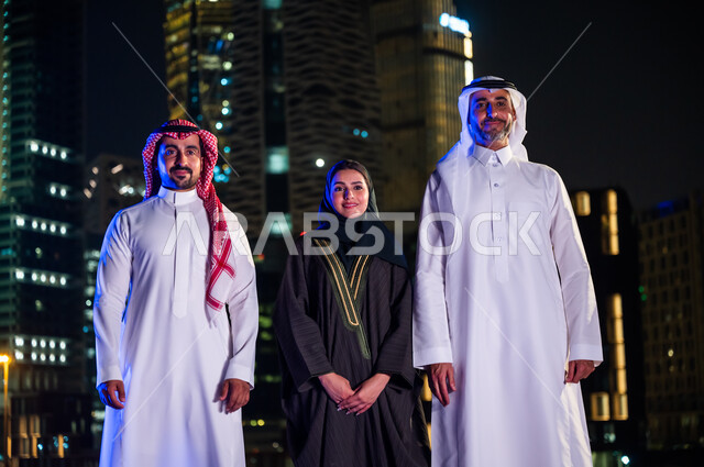 ايماءات السعادة والسرور بالتقدم والنمو, فخر أبناء المملكة بتطورها العمراني، الوقوف باستقامة وثقه واعتزاز بالدولة السعودية والنظر إلى الكاميرا، مواطنون عرب سعوديون مبتسمون يرتدون الزي التقليدي أمام أبراج وناطحات السحاب المضيئة ليلا في مدينة الرياض